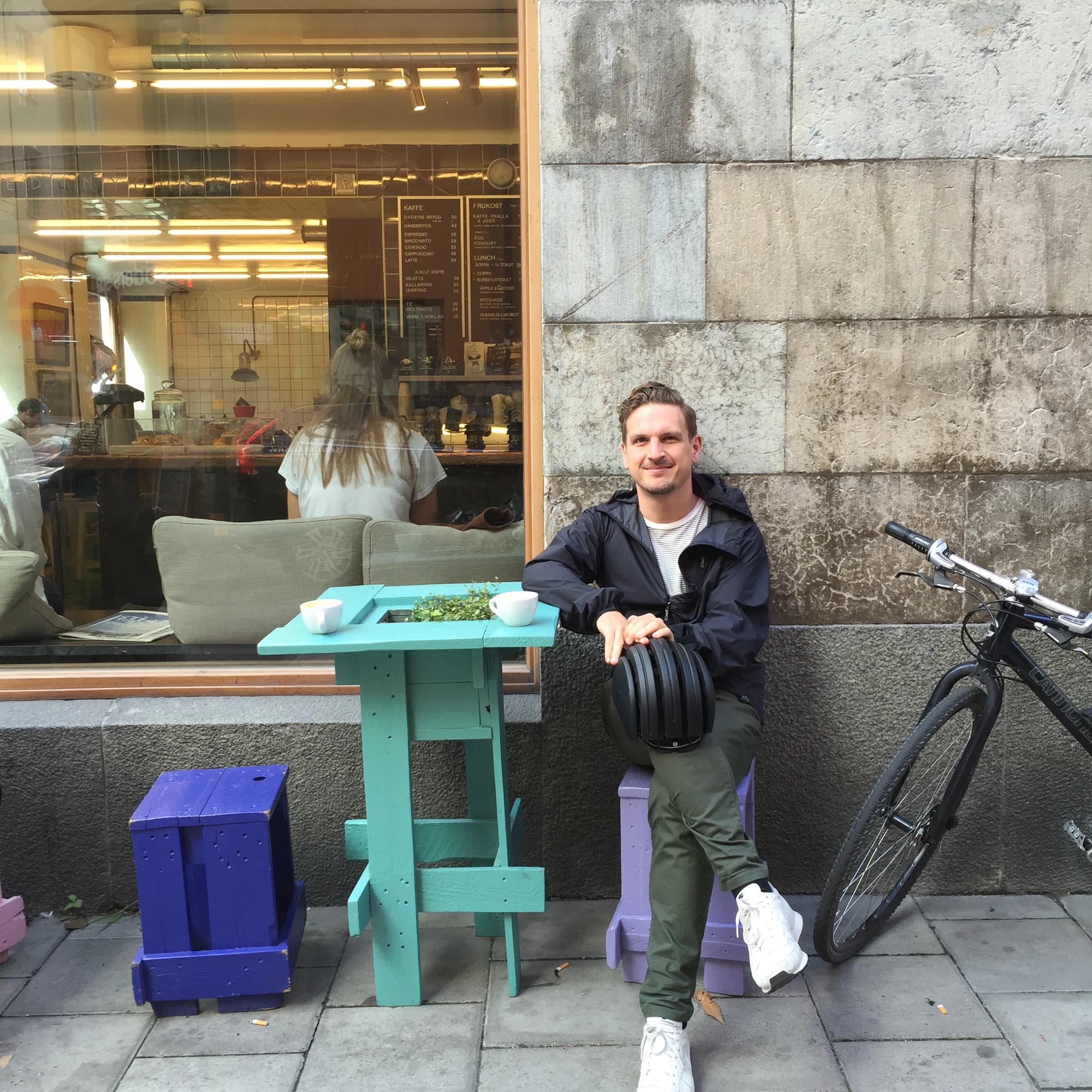 Marcus Rydling sitting outside a café on a city street, holding a bike helmet, with a bicycle beside him